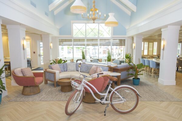 Bright, airy lounge with pastel furniture, round tables, a bicycle with a basket in the foreground, large windows, light wood flooring, potted plants, and high ceilings with modern pendant lights.