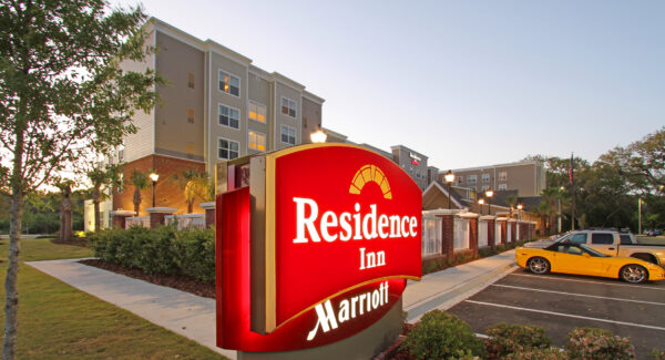 A brightly lit red and yellow Residence Inn by Marriott sign stands in front of a multi-story hotel building at sunset, with trees, parked cars, and a clear sky in the background.