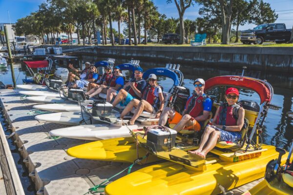 A group of people wearing life jackets sit on colorful CraigCat watercraft lined up at a dock on Amelia Island, with trees, cars, and blue skies in the background as they prepare for a group water activity.