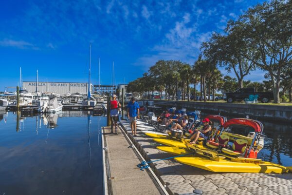 People prepare yellow water bikes at a dock beside moored boats at Amelia Island Marina, with trees and blue sky in the background. Some people stand while others sit on the water bikes.