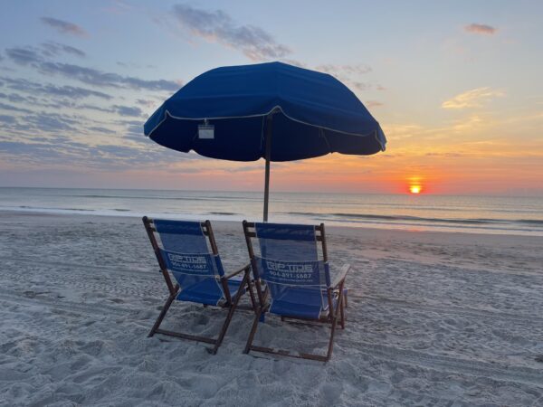 Two blue Riptide Amelia Island beach chairs sit under a large blue umbrella facing the ocean at sunrise, with the sun low on the horizon casting a warm glow over the calm water and sandy beach.