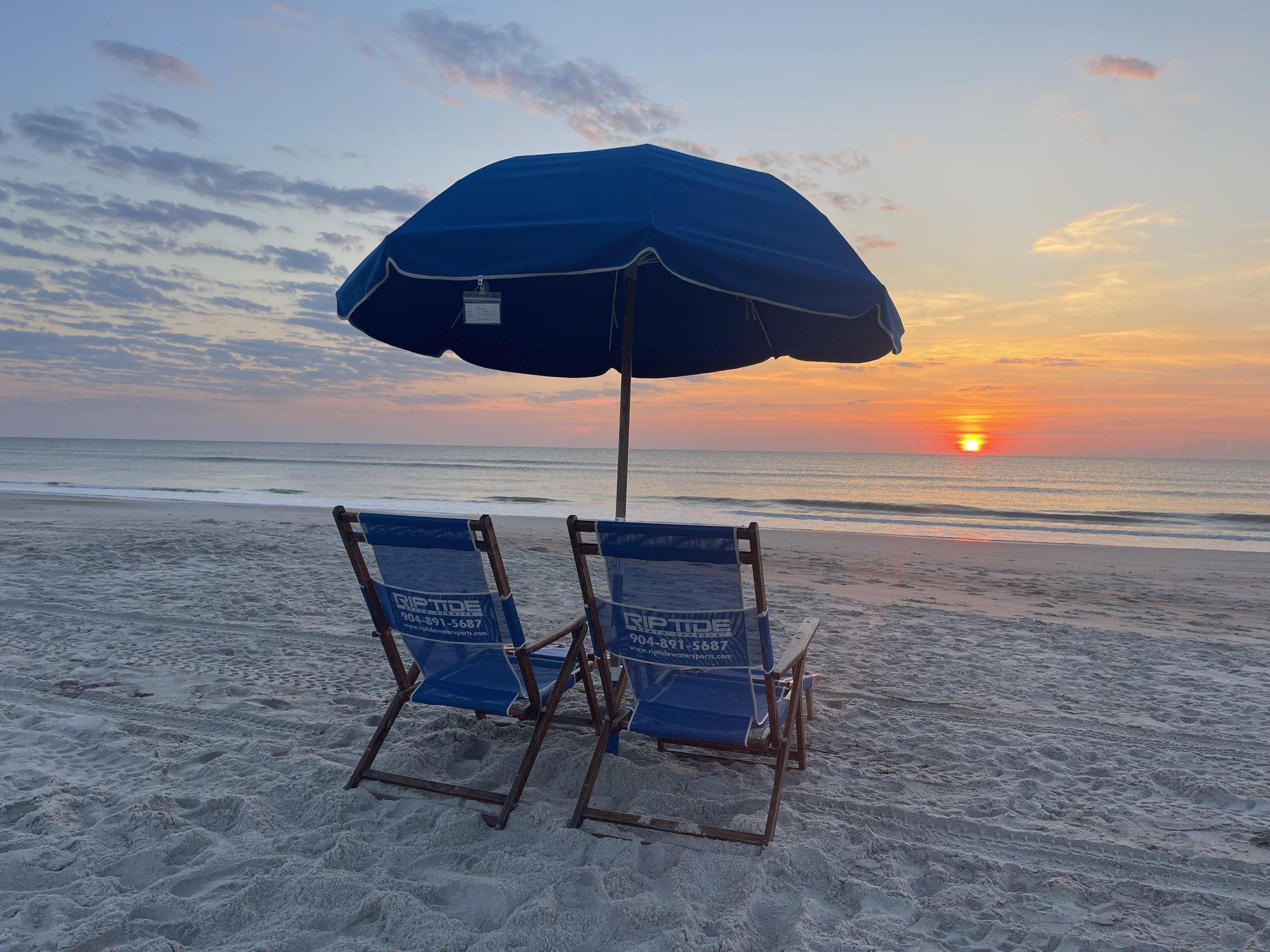 Two blue Riptide Amelia Island beach chairs sit under a large blue umbrella facing the ocean at sunrise, with the sun low on the horizon casting a warm glow over the calm water and sandy beach.
