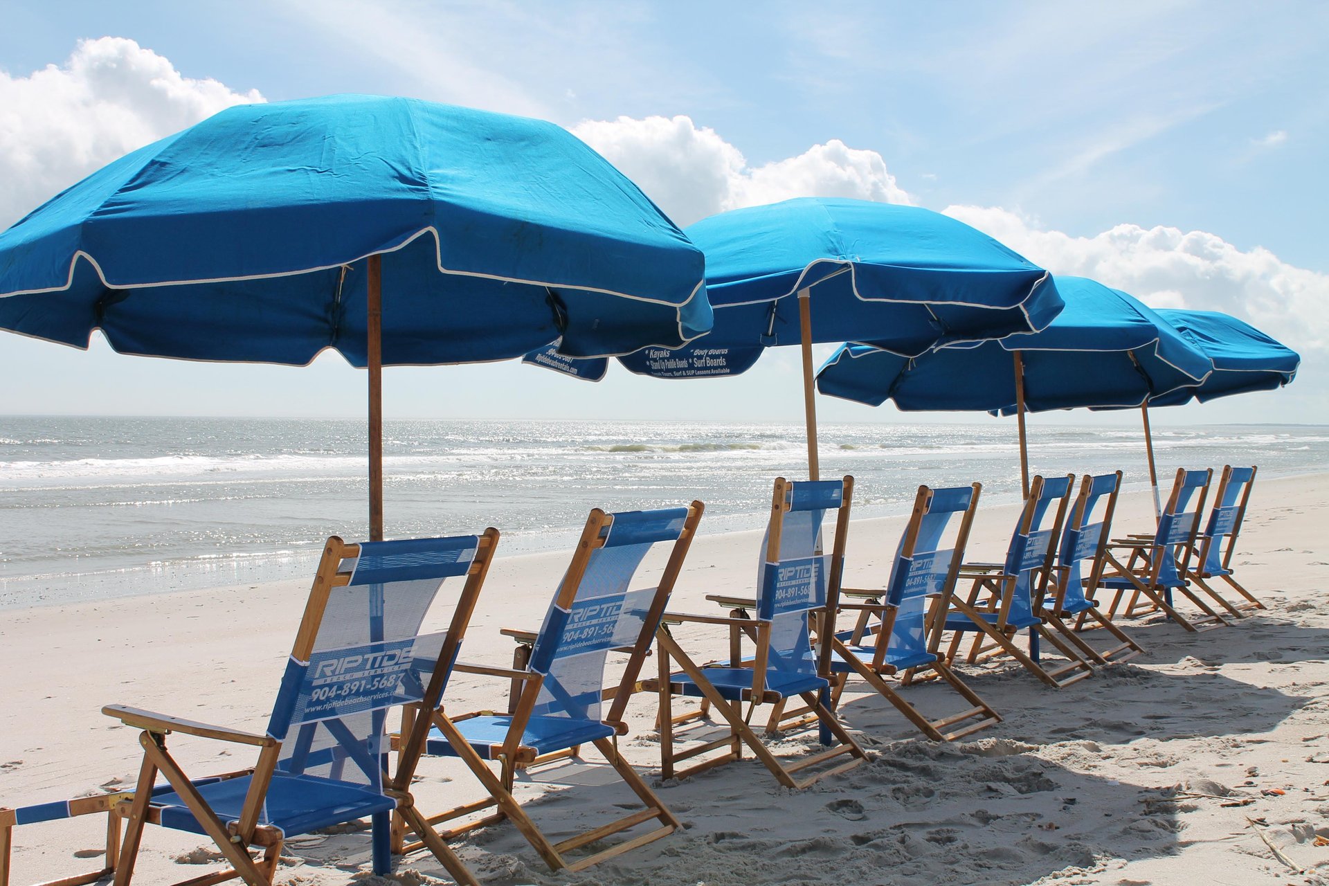 A row of empty wooden beach chairs from Riptide Amelia Island sits under blue umbrellas on the sandy shore, facing the ocean with gentle waves and a partly cloudy sky in the background.