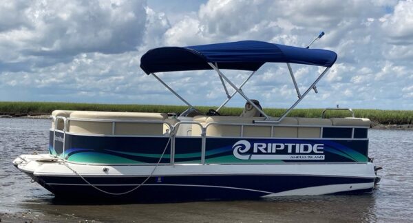 A Riptide Amelia Island pontoon boat with blue and green accents floats in shallow water near grassy marshland under a partly cloudy sky.