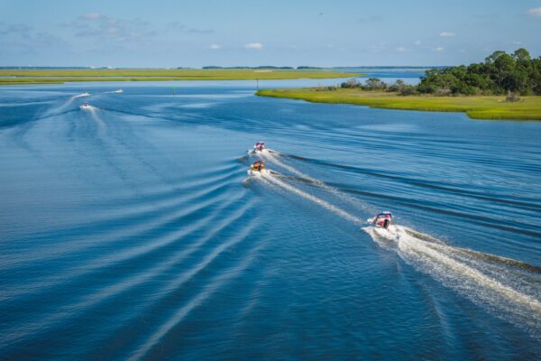 Five small boats speed across a wide, calm river, leaving curved white trails behind. The river is bordered by grassy green islands and trees under a blue sky with scattered clouds.