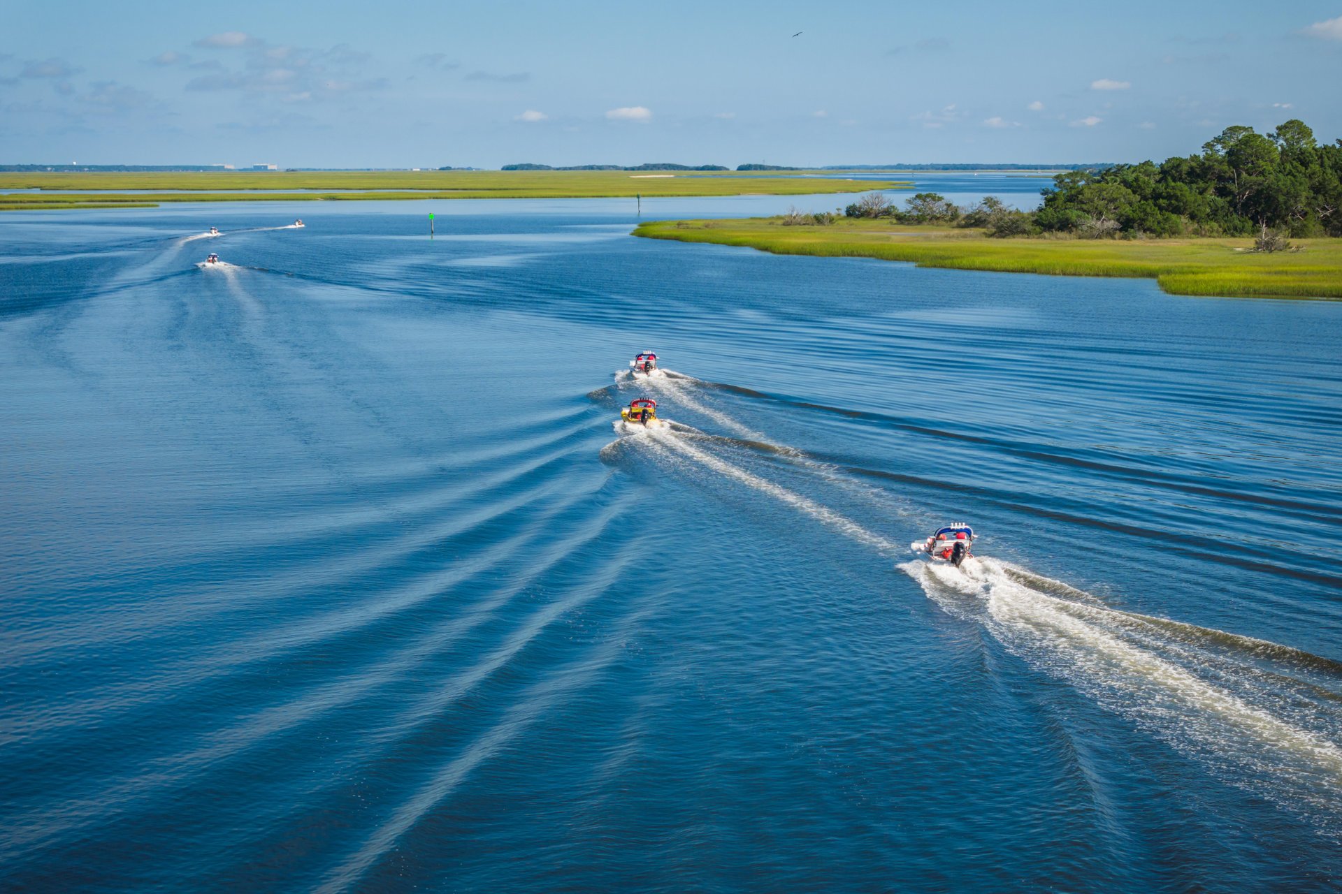 Five small boats speed across a wide, calm river, leaving curved white trails behind. The river is bordered by grassy green islands and trees under a blue sky with scattered clouds.