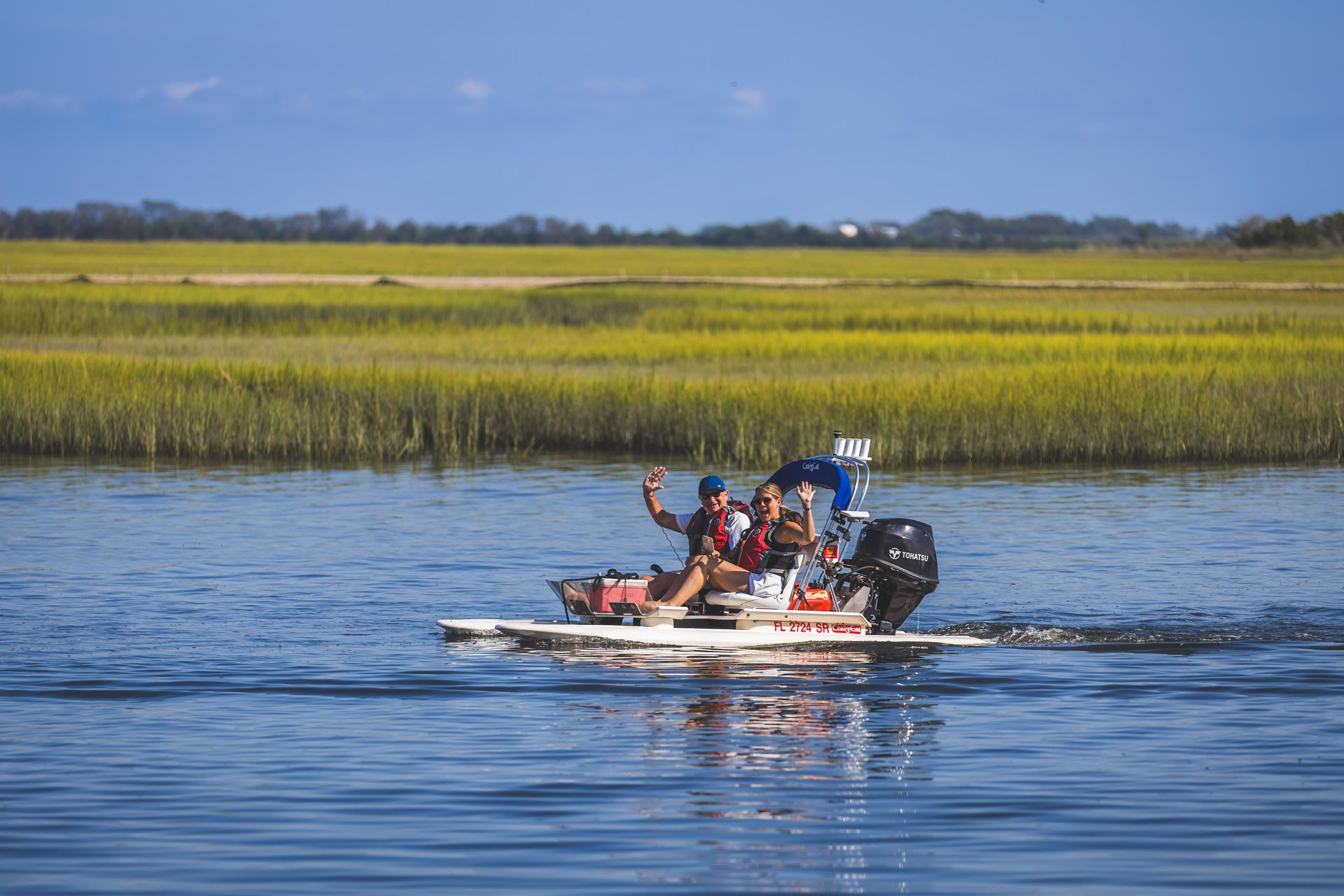 Three people ride a small motorboat on a calm river, waving and smiling. Tall green grass and distant trees line the riverbank under a clear blue sky.