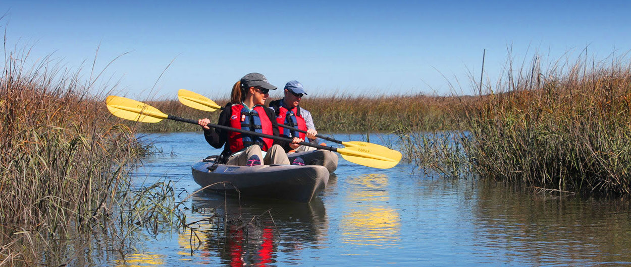 Two people wearing life jackets and hats paddle a tandem kayak with yellow oars through calm marsh waters surrounded by tall grasses under a bright blue sky.