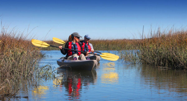 Two people wearing life jackets and hats paddle a kayak with yellow oars through calm, narrow water surrounded by tall grasses under a clear blue sky.