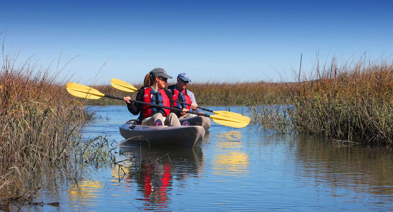 Two people wearing life jackets and hats paddle a tandem kayak with yellow oars through calm marsh waters surrounded by tall grasses under a bright blue sky.