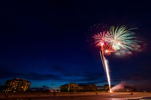 Fireworks burst in the night sky above a beach, with illuminated buildings in the background and a few people visible on the sand enjoying the display.