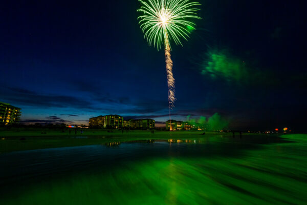 A bright green firework explodes in the night sky over a beach, illuminating the water with a green glow. Buildings are visible in the background, and the sky is dark blue with scattered clouds.