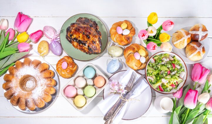 Festive served Easter dinner or brunch table, with Easter egg, tulip flower bouquet, traditional Easter foods - cake, cross buns, wreath bread, glazed ham and spring salad, white table background