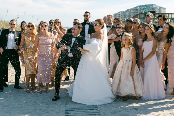 A large wedding party on a beach, with the bride and groom in the center. The groom pops a champagne bottle, spraying bubbles, while guests in formal attire smile and celebrate, with buildings visible in the background.