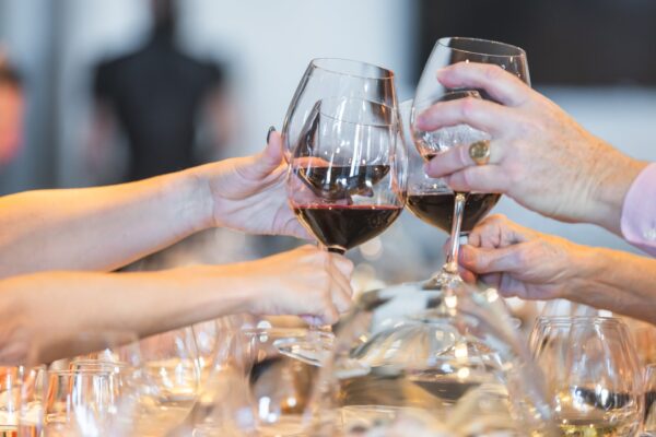 Four people clinking large glasses of red wine together in a toast, with a blurred background and a table set with more glasses and tableware.