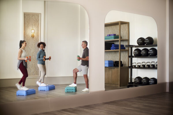 Three people exercise with dumbbells and step platforms in a bright fitness studio, smiling and facing a large mirror. Workout equipment like mats, kettlebells, and medicine balls are neatly arranged on shelves in the background.