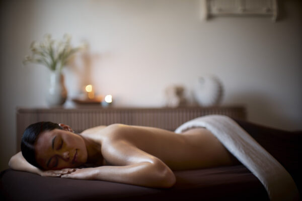 A woman is lying face down on a massage table, covered with a white towel, in a softly lit room with candles and flowers in the background. She appears relaxed and peaceful.