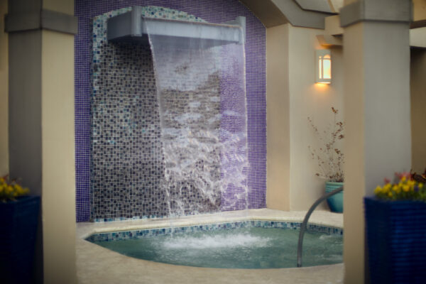 A small indoor pool with bubbling water, a curved handrail, and a waterfall feature flowing from a tiled wall with blue and purple mosaic tiles, surrounded by beige walls and soft lighting.