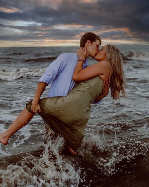 A couple kisses passionately in the ocean at sunset, with waves splashing around them. The woman lifts her leg as the man holds her waist. The sky is dramatic with clouds and warm colors.
