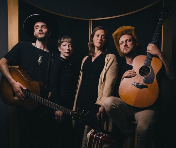 Four people pose together indoors with serious expressions; two hold acoustic guitars, one stands behind an accordion, and all wear hats or dark clothing against a dark backdrop with wooden framing.