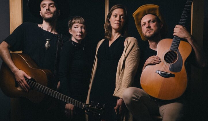 Four people pose together indoors with serious expressions; two hold acoustic guitars, one stands behind an accordion, and all wear hats or dark clothing against a dark backdrop with wooden framing.