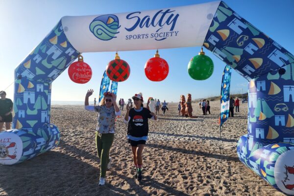Two people wearing festive hats walk under a decorated Salty Sports Society arch on a sandy beach, waving and smiling. The arch is adorned with Christmas ornaments and beach-themed designs. Others are visible in the background.