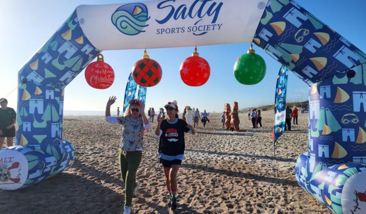 Two people wearing festive hats walk under a decorated Salty Sports Society arch on a sandy beach, waving and smiling. The arch is adorned with Christmas ornaments and beach-themed designs. Others are visible in the background.