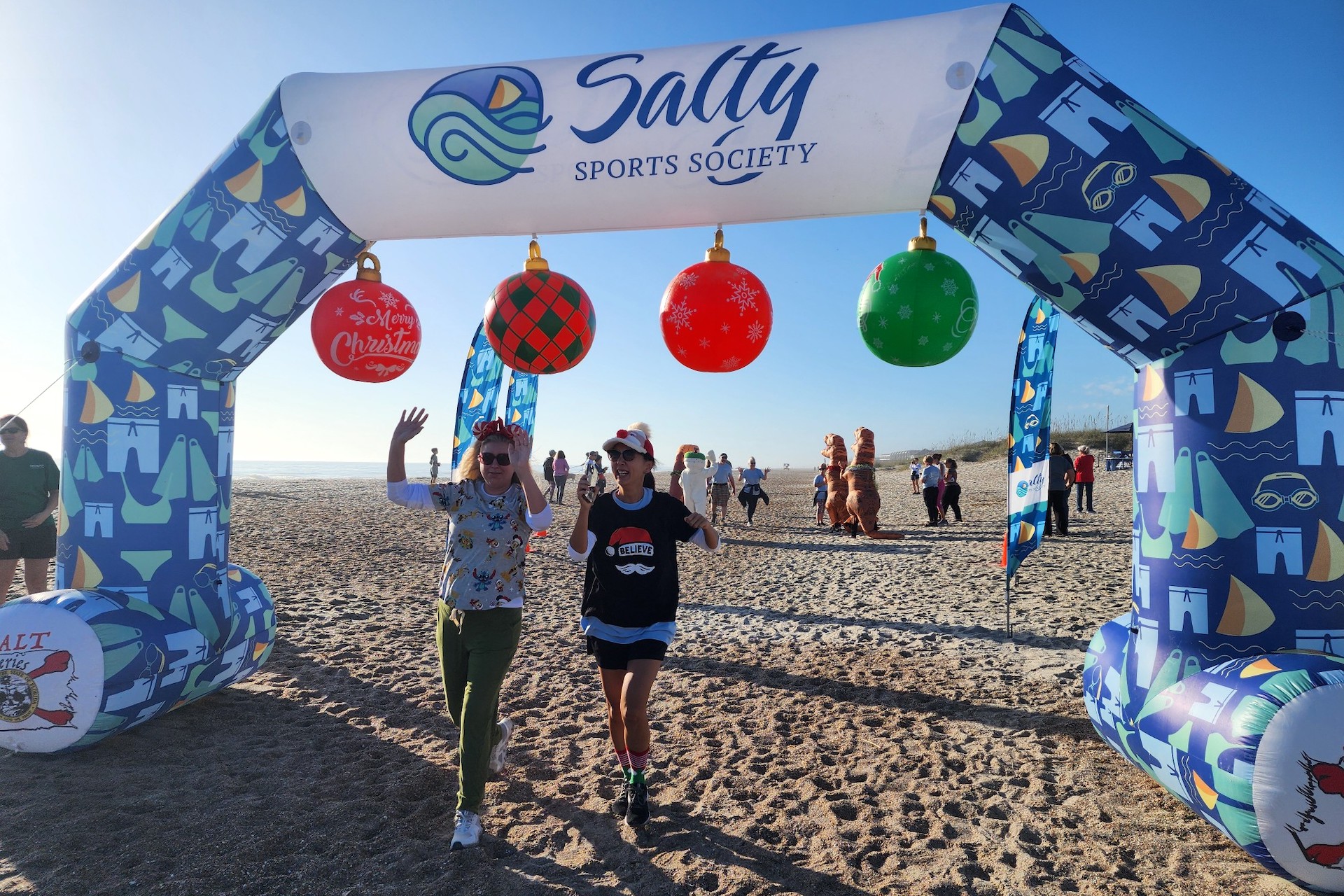 Two people wearing festive hats walk under a decorated Salty Sports Society arch on a sandy beach, waving and smiling. The arch is adorned with Christmas ornaments and beach-themed designs. Others are visible in the background.