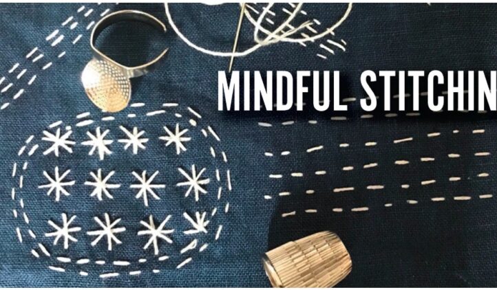 A navy blue fabric with white stitched patterns, a needle with thread, a thimble, and a metal ring. Bold text on the fabric reads “MINDFUL STITCHING.”.