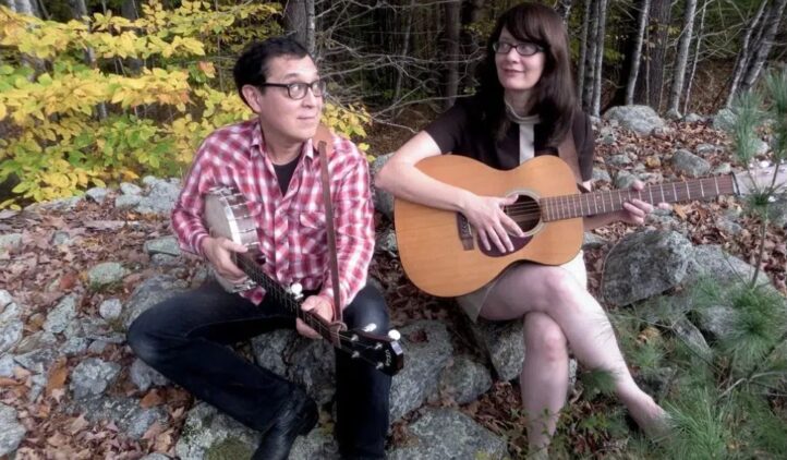 Two people sit on rocks outdoors surrounded by autumn leaves and trees, playing string instruments. The person on the left holds a banjo, and the person on the right holds an acoustic guitar. Both appear to be making music together.