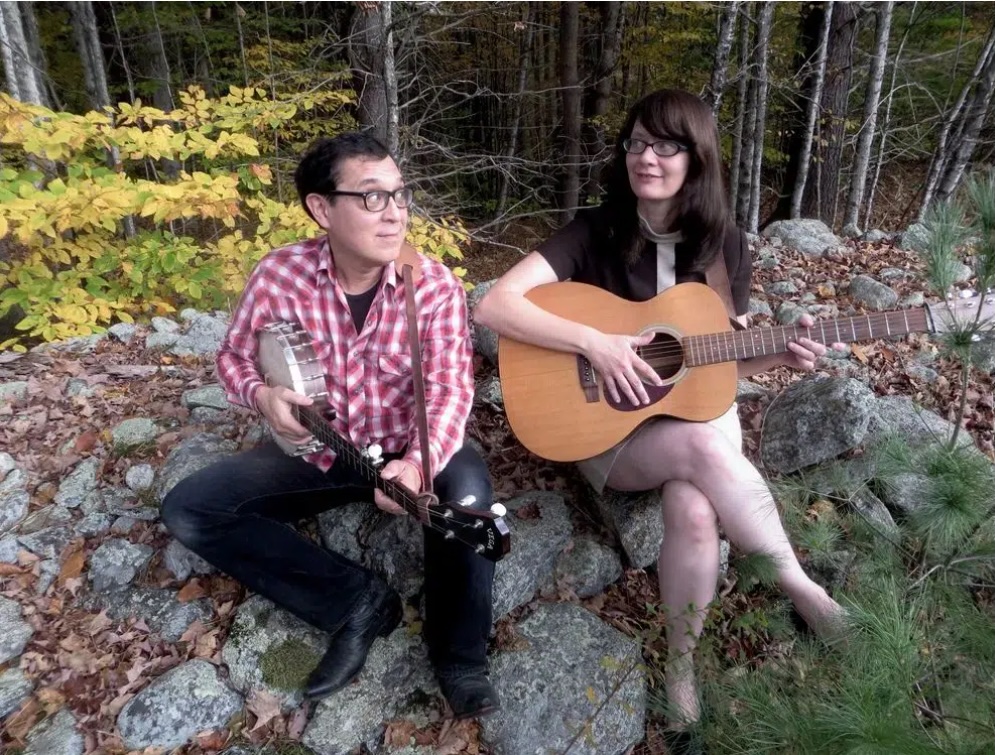 Two people sit on rocks outdoors surrounded by autumn leaves and trees, playing string instruments. The person on the left holds a banjo, and the person on the right holds an acoustic guitar. Both appear to be making music together.