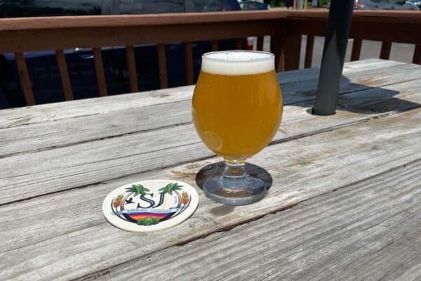 A tulip glass of hazy, golden beer sits on a weathered wooden table beside a round coaster with palm trees and SJ Brewing Company printed on it. Sunlight casts shadows across the table.