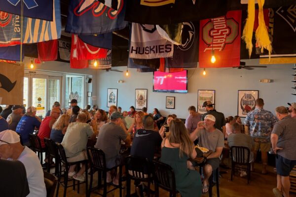 A crowded sports bar with people sitting at tables, chatting and watching a game on TV. Colorful sports flags hang from the ceiling, creating a lively atmosphere.