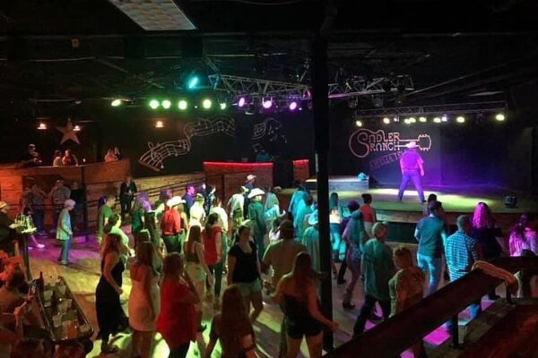 A group of people line dancing on a wooden floor under colorful lights at an indoor venue with a stage, western décor, and the Saddle Ranch sign in the background.