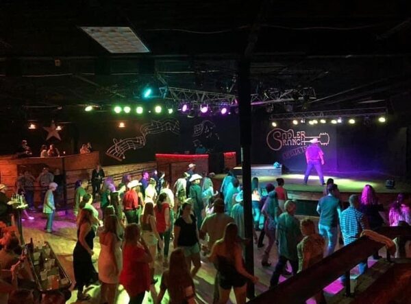 A lively crowd dances on a wooden floor under colorful lights at a western-themed venue, with music notes and Saddler Ranch painted on the wall in the background. People wear casual and cowboy-style attire.