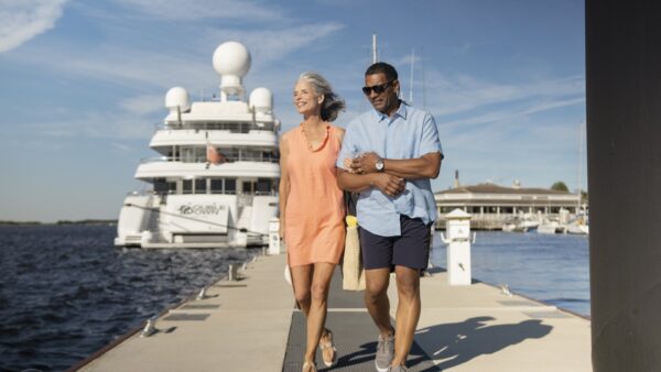 A smiling older couple walks arm in arm down a dock beside a large yacht on a sunny day. The woman wears an orange dress and carries a straw bag, while the man wears a blue shirt and sunglasses.