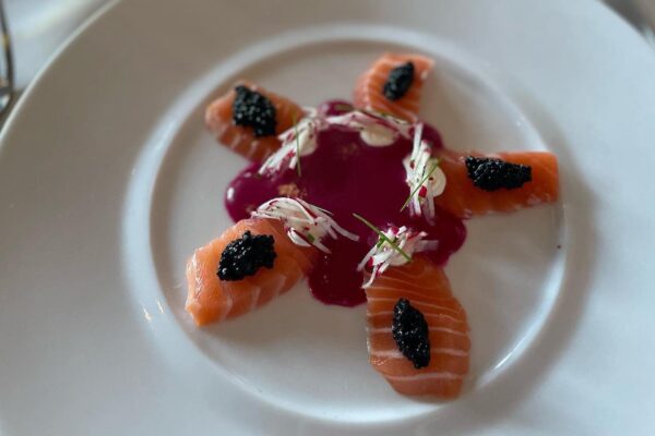 Five pieces of sliced salmon arranged in a star pattern on a white plate, topped with black caviar and thinly sliced garnish, surrounding a pool of purple sauce in the center.