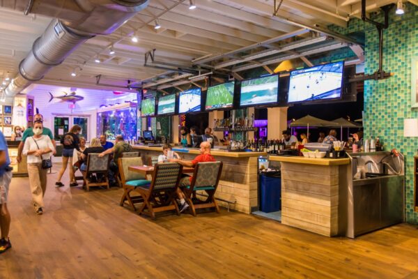 A lively bar with people sitting at tables and the counter, multiple TV screens showing sports, bright lighting, and a beach-themed decor with teal tiles and fish decorations.