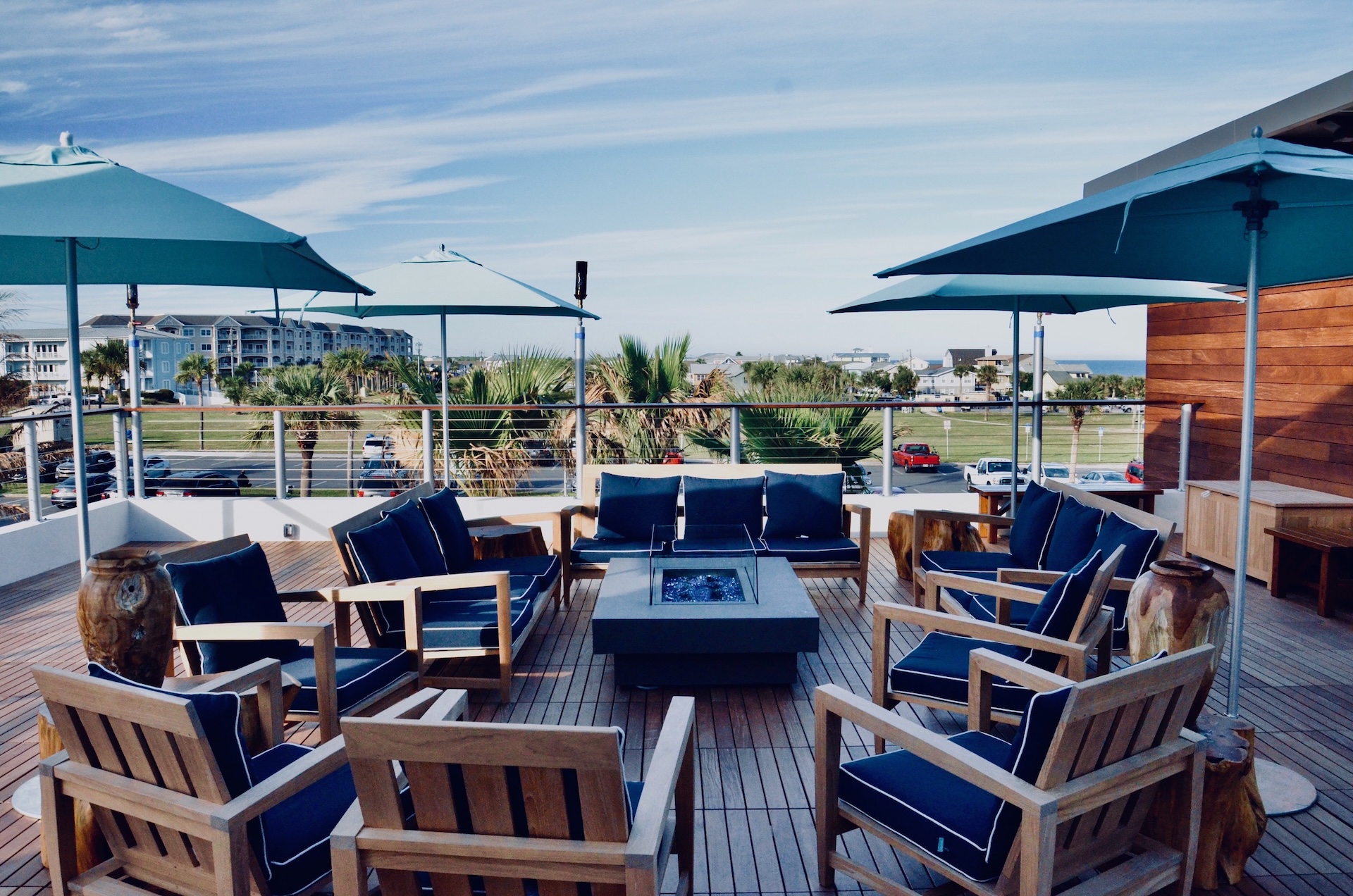 A rooftop patio with wooden chairs and blue cushions arranged around a square fire pit, shaded by teal umbrellas. Palm trees, buildings, and a blue sky with wispy clouds are visible in the background.