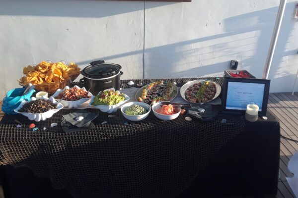 A buffet table is set with tortilla chips, bowls of salsa, guacamole, black beans, and various toppings. There is also a slow cooker, plates of sushi, decorative seashells, and a framed sign on a black tablecloth.