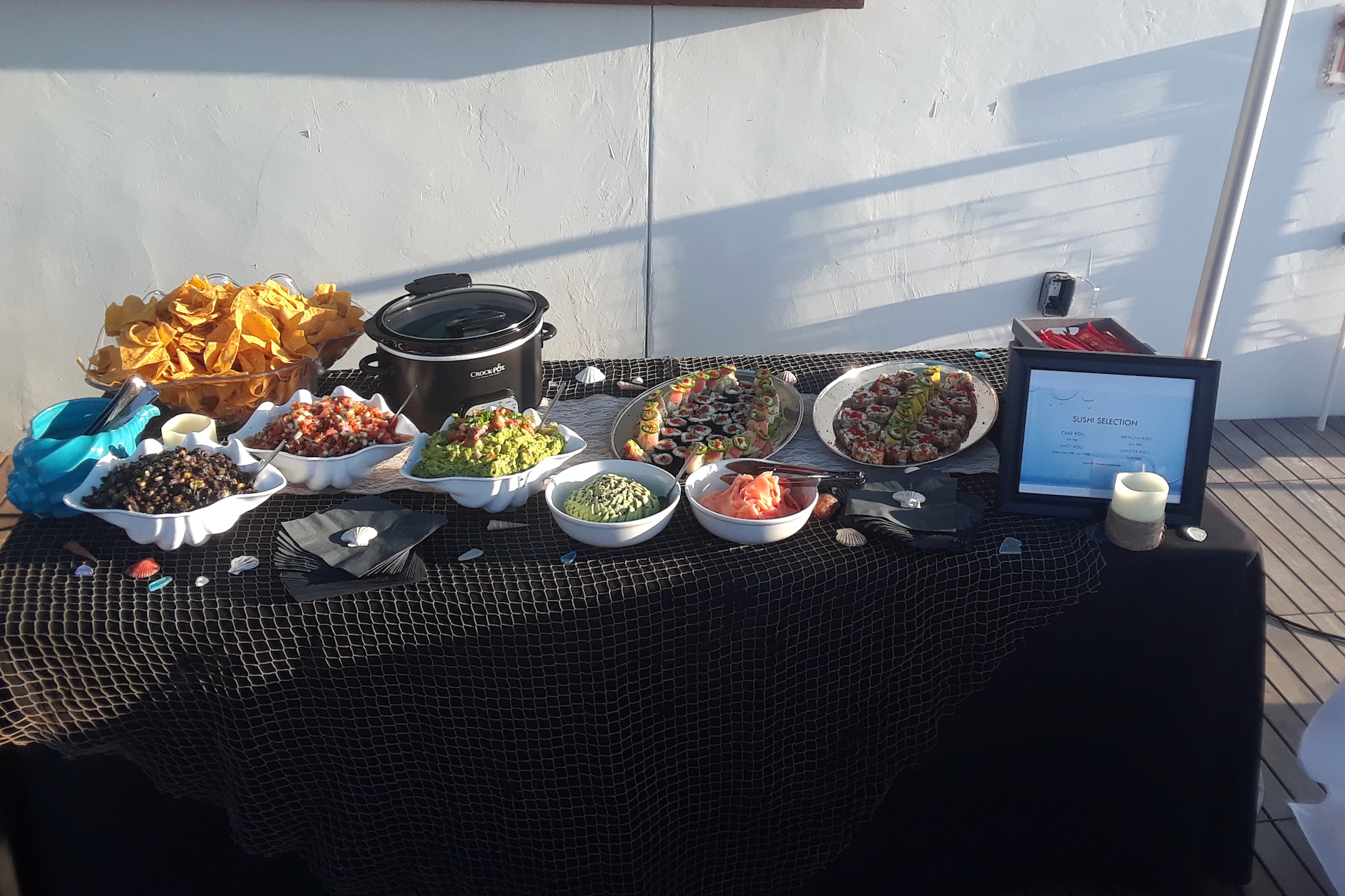 A buffet table is set with tortilla chips, bowls of salsa, guacamole, black beans, and various toppings. There is also a slow cooker, plates of sushi, decorative seashells, and a framed sign on a black tablecloth.