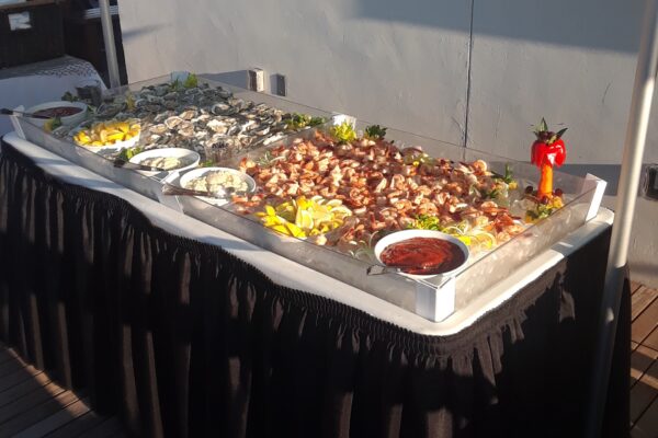 A seafood buffet on ice with shrimp, oysters, lemon wedges, cocktail sauce, and garnishes, displayed on a table with a black tablecloth against a gray wall in sunlight.