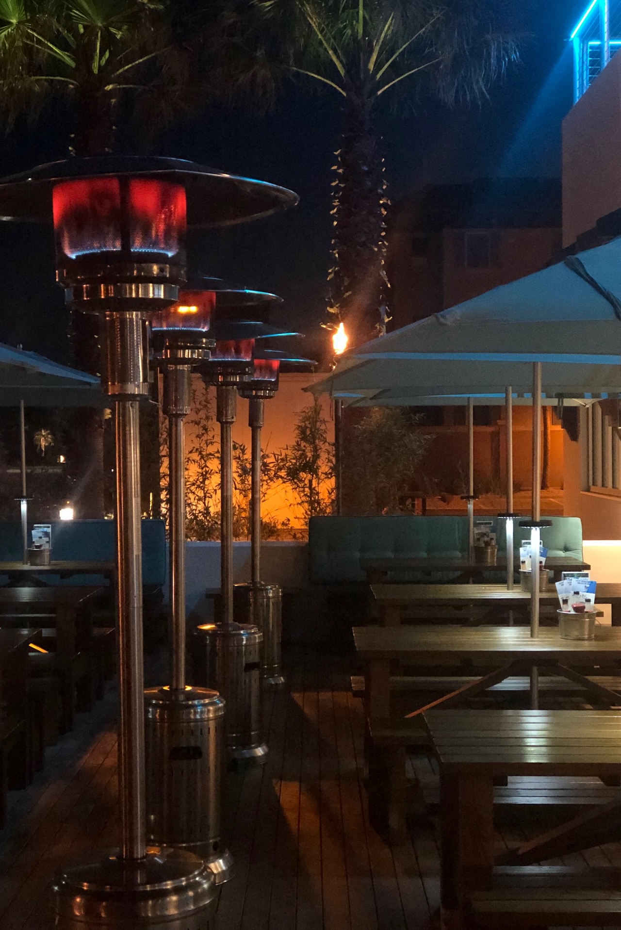 Outdoor restaurant seating at night with wooden tables, lit patio heaters, large blue umbrellas, and palm trees in the background, creating a cozy, inviting atmosphere.