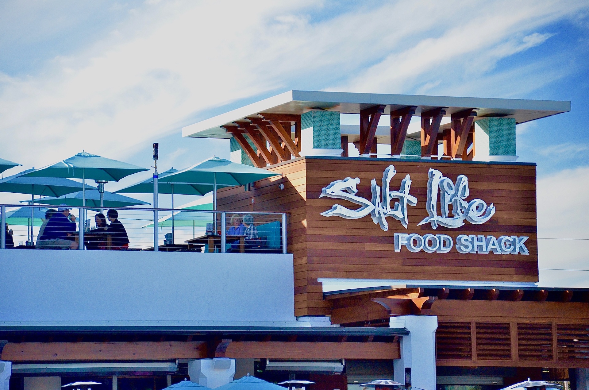 A modern beachside restaurant with a sign reading Salt Life Food Shack features outdoor seating and blue umbrellas under a partly cloudy sky.