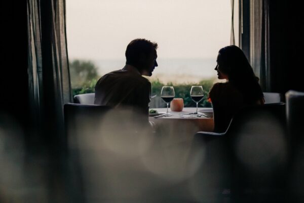 A man and a woman sit across from each other at a table set for two by a window, softly lit, with wine glasses and a scenic view outside, creating a romantic and intimate atmosphere.