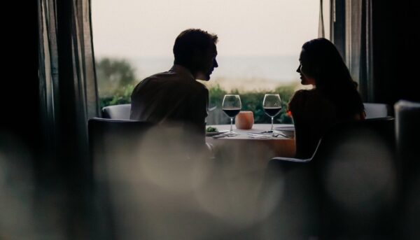 A man and woman sit facing each other at a table by a window, sharing a romantic dinner with wine glasses, softly lit by natural light from outside.
