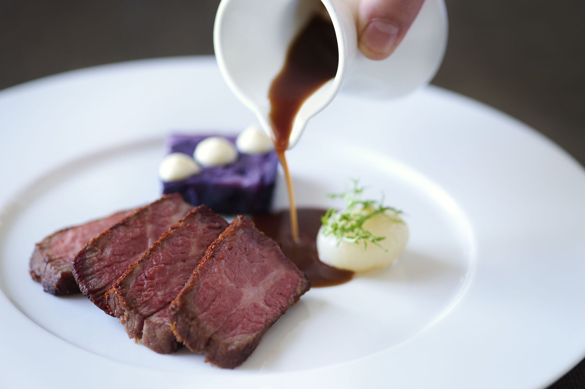 Slices of seared beef on a white plate, with sauce being poured from a small pitcher. The dish is garnished with a cube of purple potato topped with cream dots and a small white vegetable with greens.