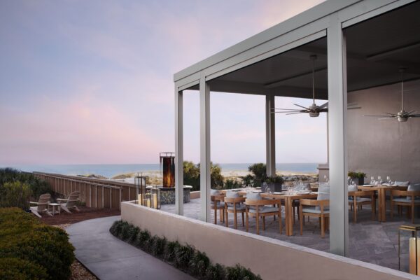 An elegant outdoor dining area with wooden tables and chairs sits under a modern pergola, overlooking sand dunes and the ocean at sunset. A fire feature and lounge chairs are visible in the background.