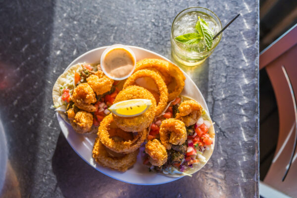 A plate of fried shrimp and crispy onion rings, served with coleslaw, pico de gallo, a lemon wedge, and dipping sauce. A glass of iced drink with mint garnish is beside the plate on a metallic table.