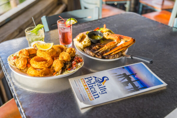 Two plates of seafood, including fried shrimp, onion rings, and crab legs, sit on a table with two cocktails. A Salty Pelican menu rests nearby. The setting appears to be a casual, sunlit restaurant.